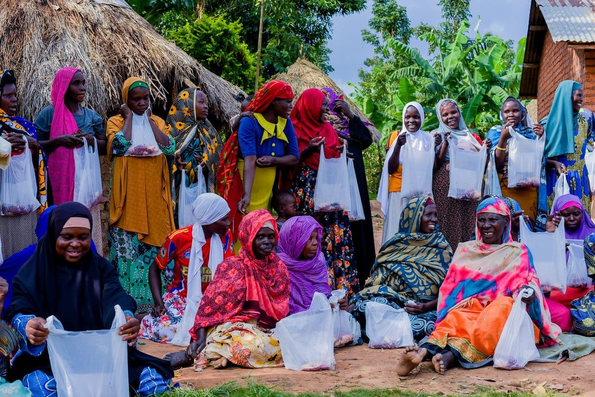 A group of women seated and standing outside a thatched home, each holding a parcel of Qurbani meat