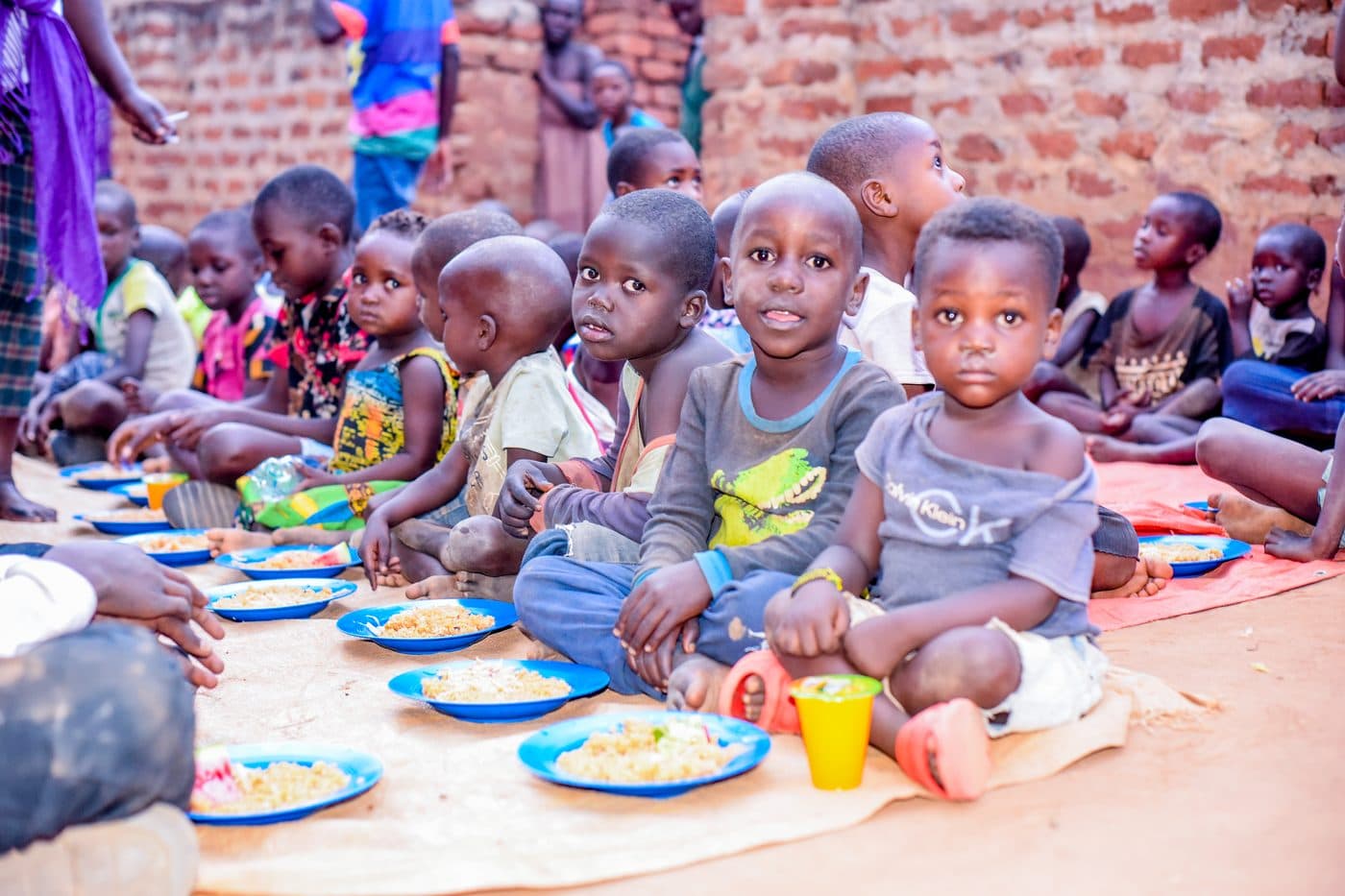 Young children sitting in a row on the ground, each with a blue plate of rice in front of them, waiting for the daily hot meal to begin