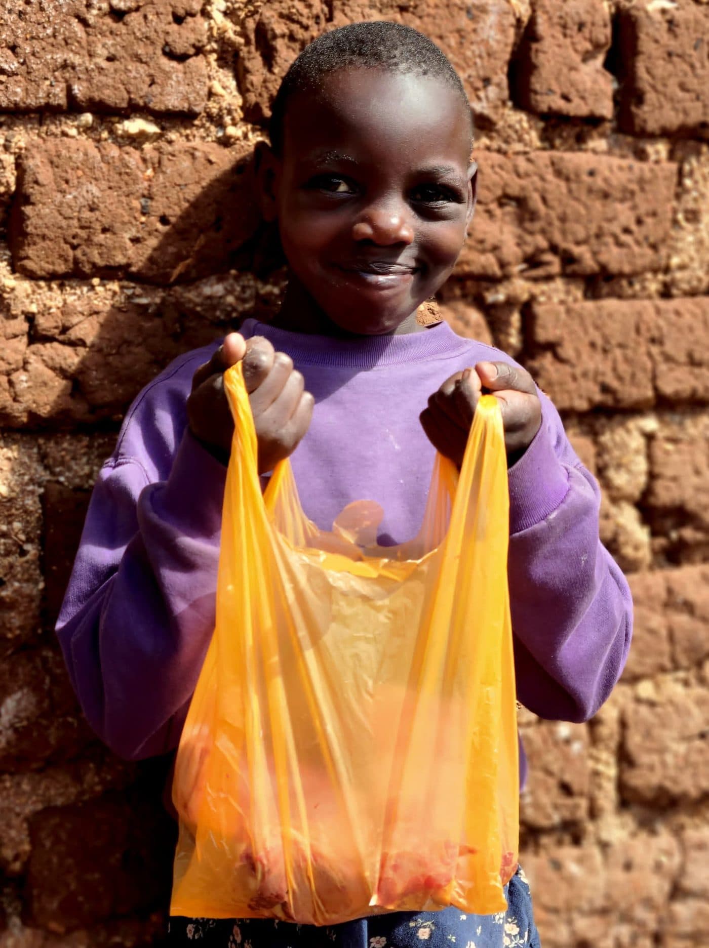 A smiling child holding up an orange bag of Qurbani meat outside a brick home