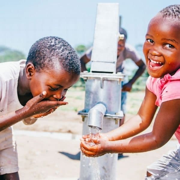 Two children laughing at a borehole pump as clean water runs over their hands
