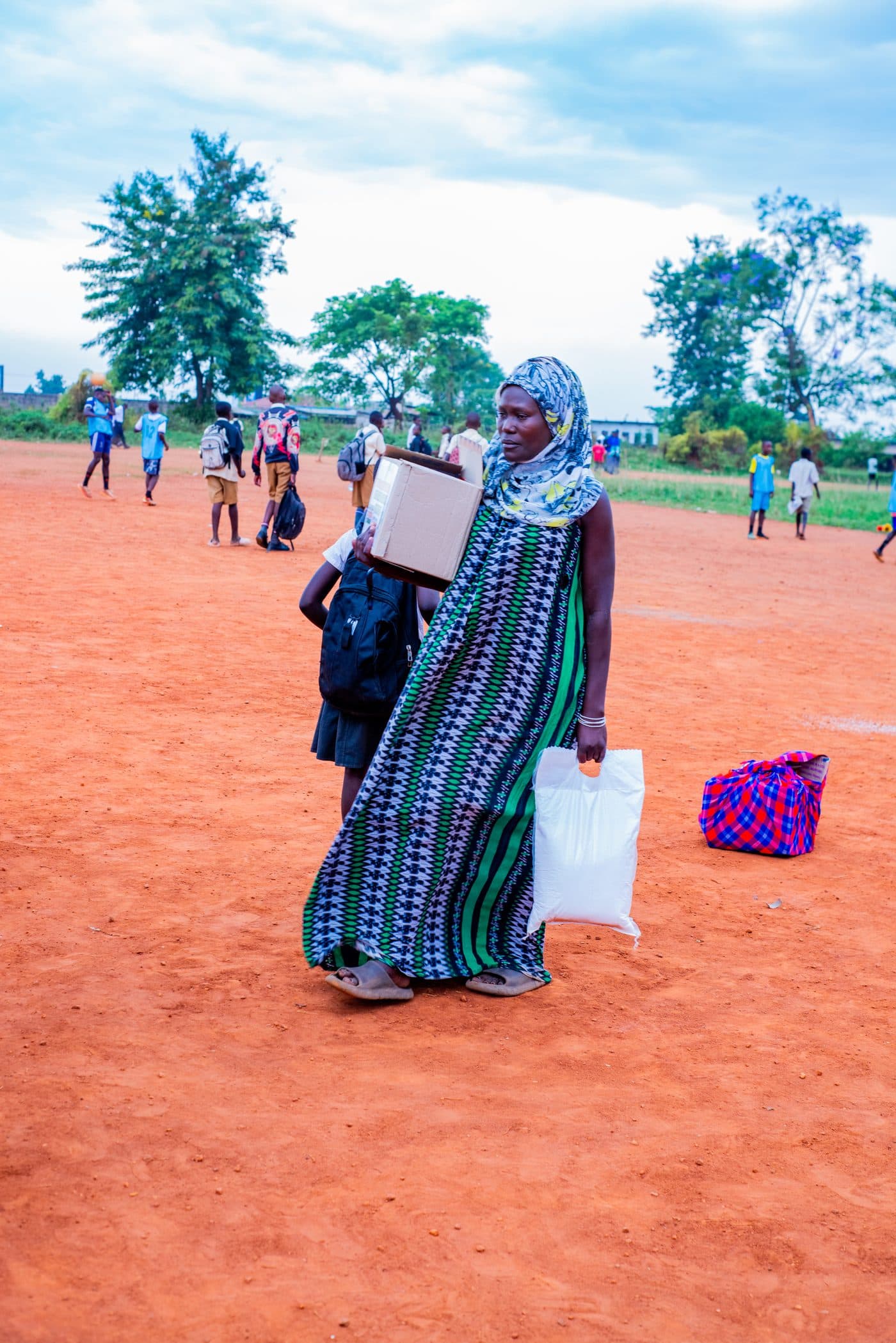 A woman carrying a box and a sack of food home across a field after a distribution