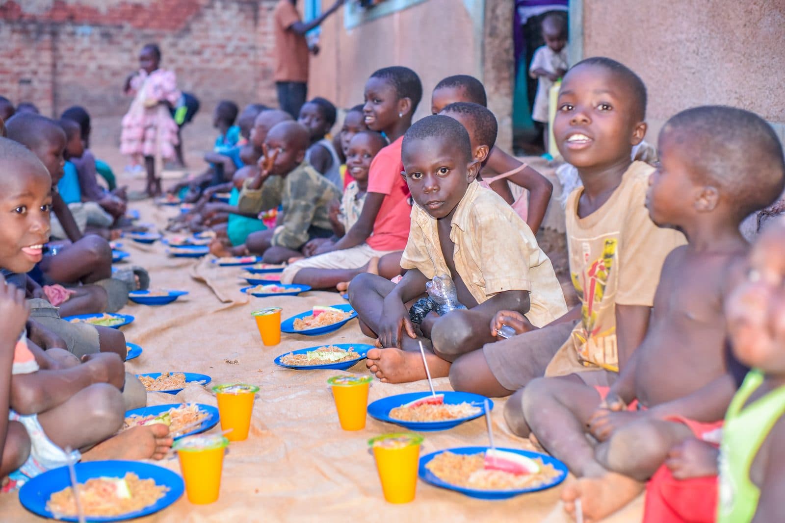 Children seated along a row of blue plates of food at a daily feeding, several looking up at the camera